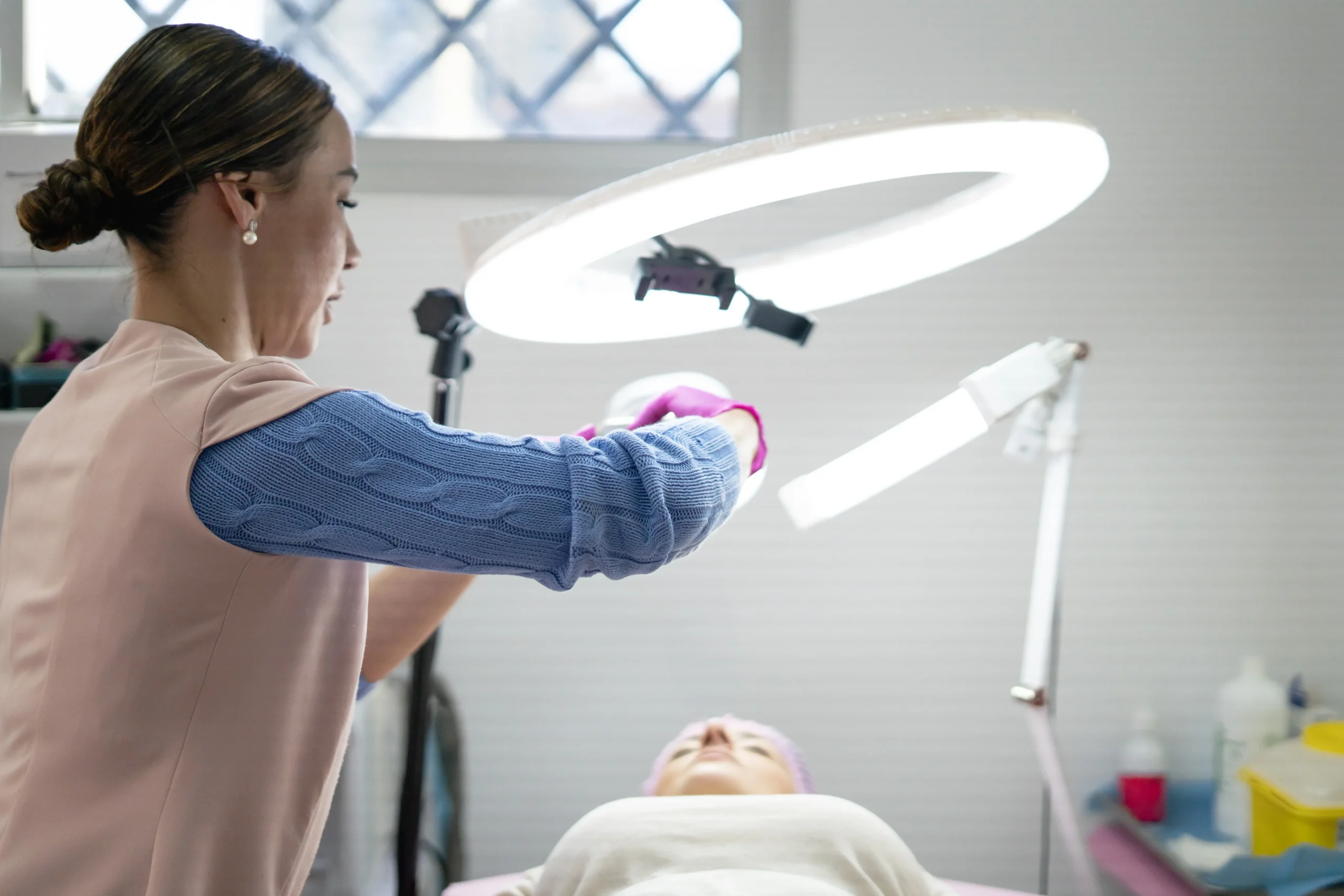 Aesthetic professional adjusting lighting equipment above a patient during a facial or laser procedure in a clinical treatment room.